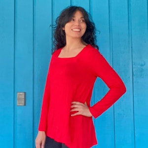 May include: A woman wearing a red long-sleeved top with a square neckline. The top has a slightly A-line shape and is made of a soft, stretchy fabric. She is standing in front of a blue wall.