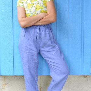 May include: A woman wearing a yellow and white floral print top with a blue linen pants and brown ankle boots. She is standing in front of a blue wall.