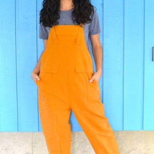 Könnte beinhalten: Eine Frau in einem blauen T-Shirt und einer orangefarbenen Latzhose steht vor einer blauen Wand. Sie lächelt und schaut in die Kamera.