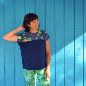 May include: A navy blue t-shirt with a floral pattern across the top and sleeves. The shirt has a color block design with a solid navy blue section. The model is wearing green and white patterned pants.