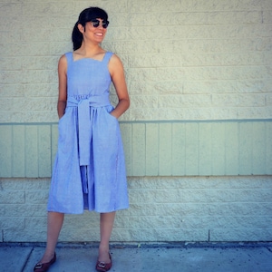May include: A woman wearing a blue and white gingham dress with a red trim. The dress has a fitted bodice and a full skirt. She is wearing sunglasses and has her hair pulled back.