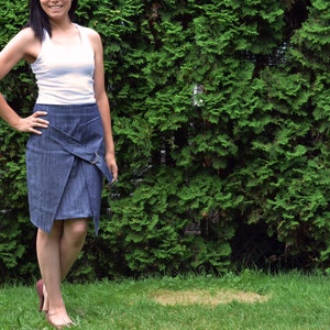 May include: A woman wearing a white tank top and a denim wrap skirt. She is standing in front of a green hedge.