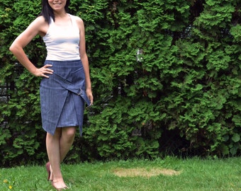 May include: A woman wearing a white tank top and a denim wrap skirt. She is standing in front of a green hedge.
