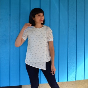 May include: A woman wearing a white blouse with a small bicycle print and dark blue jeans. The blouse has a unique asymmetrical hemline.