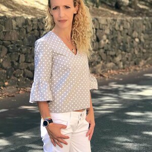 May include: A woman wearing a gray and white polka dot blouse with a ruffled sleeve and white pants. She is wearing a watch on her left wrist.