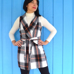 May include: A white, black, and orange plaid vest with a shawl collar and a self-tie belt. The vest is worn over a white turtleneck.