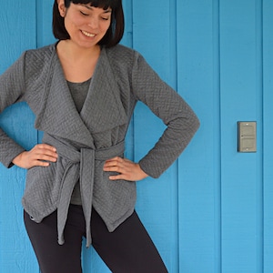 Könnte beinhalten: Eine graue gesteppte Wickeljacke mit einem langen, spitzen Kragen und einem Bindegürtel. Die Jacke wird von einer Person getragen, die vor einer blauen Wand steht.