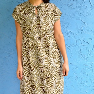 Può includere: Una donna indossa un vestito a stampa foglie verde e bianco con un collo a cravatta. Il vestito ha maniche corte e una vestibilità ampia.