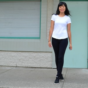 Puede incluir: Una mujer con una camiseta blanca y pantalones negros camina frente a un edificio con una puerta verde.