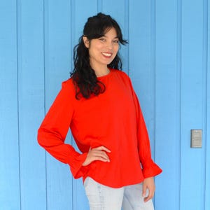 May include: A woman wearing a red long-sleeved blouse with ruffled cuffs. The blouse has a round neckline and is made of a soft, flowing fabric. She is standing in front of a blue wall.