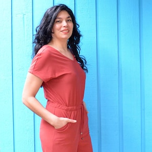 May include: A woman wearing a rust-coloured jumpsuit with a v-neck and short sleeves. The jumpsuit has a drawstring waist and pockets. She is standing in front of a blue wall.