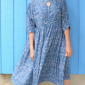 May include: A woman wearing a blue and white polka dot dress with a keyhole neckline. She is standing in front of a blue wall and wearing brown ankle boots.