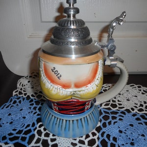 May include: A ceramic beer stein with a silver lid and handle. The stein is decorated with a woman in a traditional German dirndl dress. The year 2012 is written on the front of the stein.