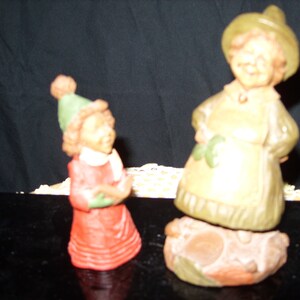May include: Two ceramic figurines. One is a child in a red dress and green hat. The other is an older woman in a yellow dress and hat. The figurines are painted with muted colors and have a vintage aesthetic.