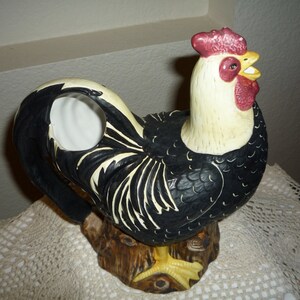 Peut inclure: Figurine de coq noir et blanc avec une cr&ecirc;te et des barbillons rouges. Le coq est debout sur une souche d'arbre brune. La figurine est en c&eacute;ramique et a un trou dans le dos pour servir de vase.