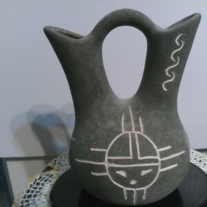May include: A gray ceramic vase with a traditional Native American design. The vase has two spouts and a handle. The design features a stylized sun symbol with a face in the center.