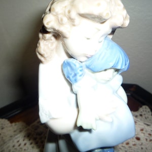May include: A porcelain figurine of a girl holding a smaller girl doll. The larger girl has blonde hair and is wearing a white dress with blue trim. The doll has blue hair and is wearing a blue dress.