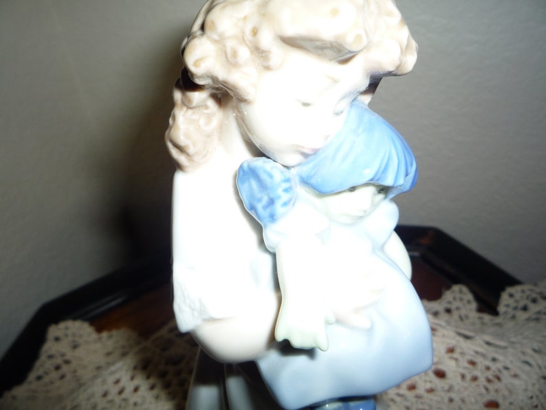 May include: A porcelain figurine of a girl holding a smaller girl doll. The larger girl has blonde hair and is wearing a white dress with blue trim. The doll has blue hair and is wearing a blue dress.