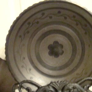 May include: A black ceramic bowl with a decorative design of stylized flowers and vines. The bowl has a concentric circle pattern and a raised rim.