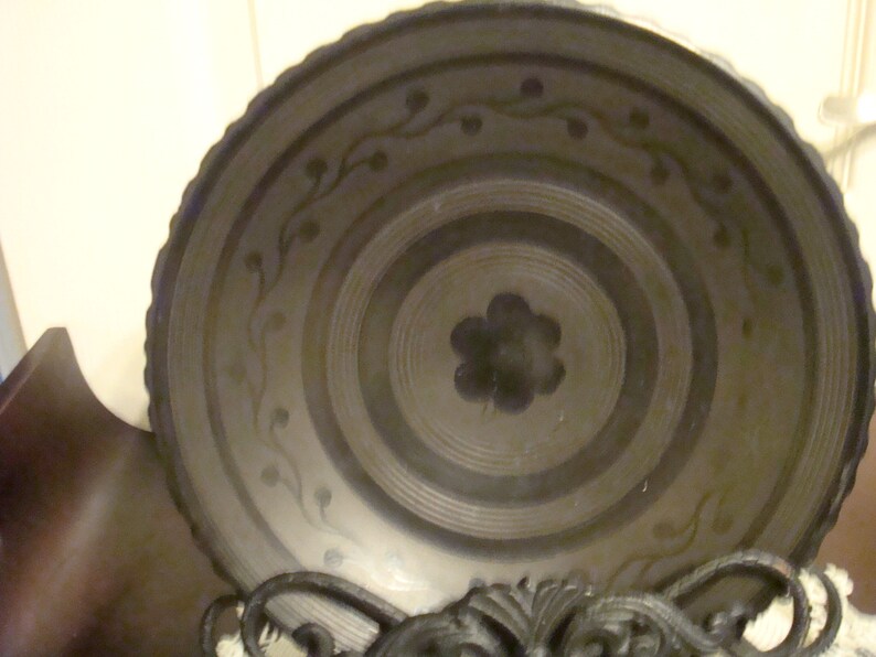 May include: A black ceramic bowl with a decorative design of stylized flowers and vines. The bowl has a concentric circle pattern and a raised rim.