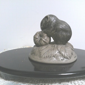 May include: A detailed, gray ceramic sculpture of a small animal, possibly a beaver, holding a pumpkin. The sculpture rests on a base with leaf details. The base sits on a black, round surface.