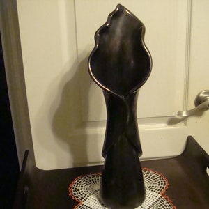 May include: A tall, dark brown ceramic vase with a calla lily shape. The vase has a flared opening and a slender, elongated body. It is placed on a dark brown tray with a white and orange doily.
