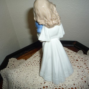 May include: A porcelain figurine of a girl in a white dress holding a blue bird. The figurine is standing on a white crocheted doily.