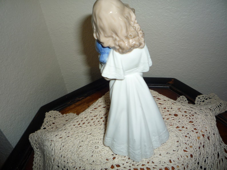 May include: A porcelain figurine of a girl in a white dress holding a blue bird. The figurine is standing on a white crocheted doily.