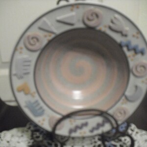 May include: A white ceramic bowl with a light blue and pink swirl design. The bowl has a decorative rim with various shapes and colors.