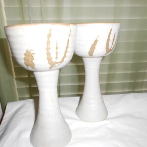 May include: Two white ceramic cups with brown designs. The cups are on a white surface.