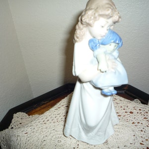 May include: A porcelain figurine of a girl holding a smaller porcelain doll. The girl is wearing a white dress and has long brown hair. The doll is wearing a blue dress and has blue hair.