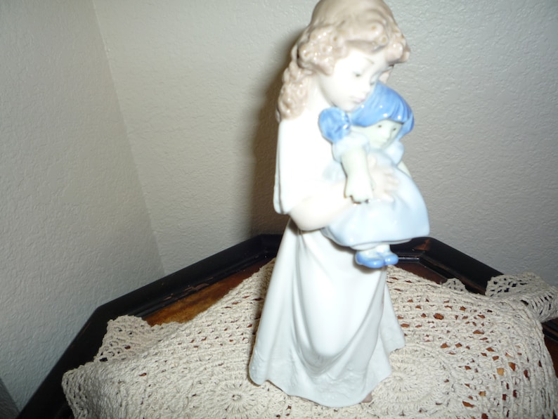 May include: A porcelain figurine of a girl holding a smaller porcelain doll. The girl is wearing a white dress and has long brown hair. The doll is wearing a blue dress and has blue hair.