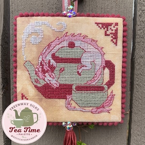 May include: A cross-stitch embroidery of a teapot with a pink dragon wrapped around it. The embroidery is framed with a red pom-pom trim. The text "Tea Time" is on a pink circle with a smaller teapot image. The text "Treeway Silks - Palette" is above the circle. The text "@xstitchtherainbow" is below the circle.