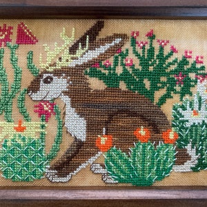 Puede incluir: Un conejo marrón y blanco con una corona de cuernos está bordado sobre un fondo beige. El conejo está rodeado de plantas de cactus verdes con flores rosas y blancas.