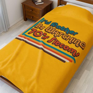 Peut inclure: Une couverture de lit jaune moutarde avec l'inscription "I'd Rather Be Watching 70's Sitcoms" dans une police rétro. Le motif comprend une bordure rayée bleue, orange, marron et blanche. La couverture est sur un cadre de lit en bois.