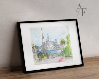 May include: Watercolor painting of Space Mountain in a black frame. The artwork depicts the iconic attraction with a white and gray exterior, set against a blue sky. Palm trees and greenery add color to the scene. The print is on a wooden surface.