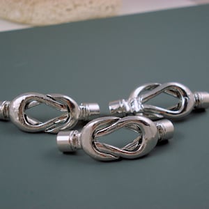 May include: Three silver-colored metal decorative knots. Each knot is in the shape of a figure eight, with a cylindrical end on each side. The knots are arranged on a gray surface.