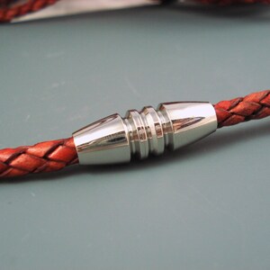 May include: Close-up of a braided leather cord with a silver-colored metal clasp. The leather is a reddish-brown color, and the clasp has a cylindrical design with ridged detailing. The background is a solid gray.