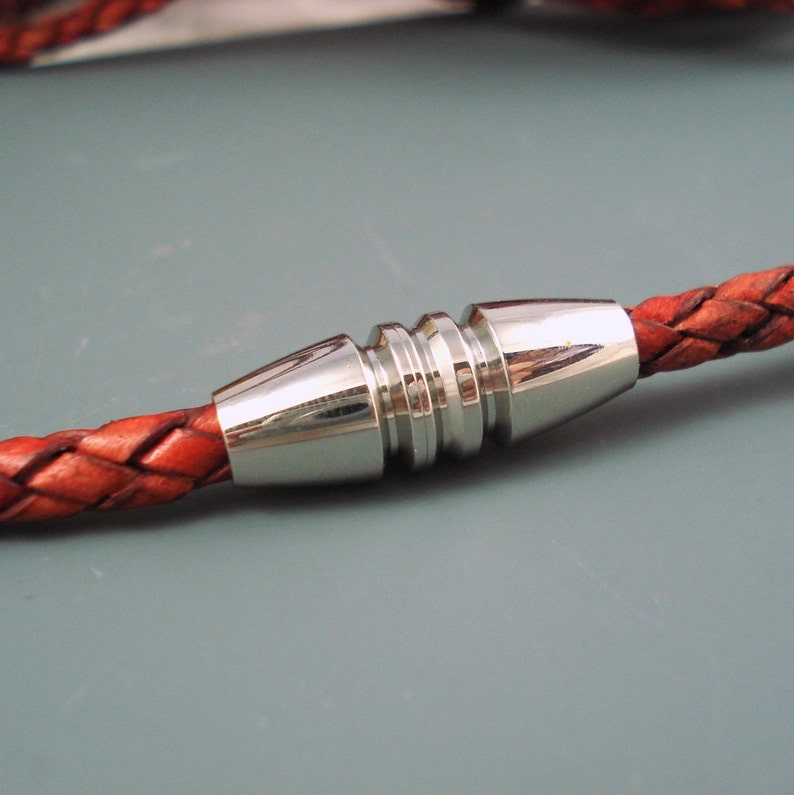May include: Close-up of a braided leather cord with a silver-colored metal clasp. The leather is a reddish-brown color, and the clasp has a cylindrical design with ridged detailing. The background is a solid gray.