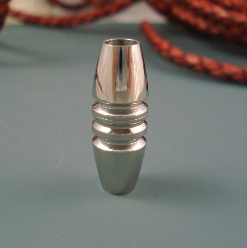 May include: A polished silver metal bead with a conical shape. The bead has a cylindrical top and is accented with three horizontal ridges. The background is a muted teal color with a blurred red rope.