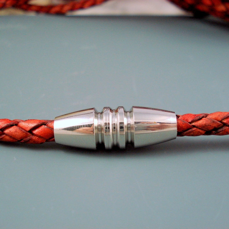 May include: Close-up of a braided leather bracelet with a silver magnetic clasp. The bracelet is a reddish-orange color, and the clasp is a shiny silver. The clasp has a ridged design in the center.