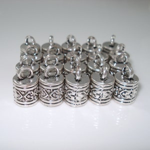 May include: A group of 18 silver metal cord end caps with a decorative design. The caps have a small loop at the top for attaching to a cord.