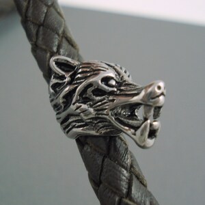 May include: A silver wolf head charm with a braided leather bracelet. The wolf's mouth is open and its teeth are visible.