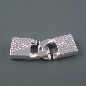 May include: Silver-toned metal clasp with a decorative design. The clasp is made of two pieces that fit together to secure a chain or bracelet.