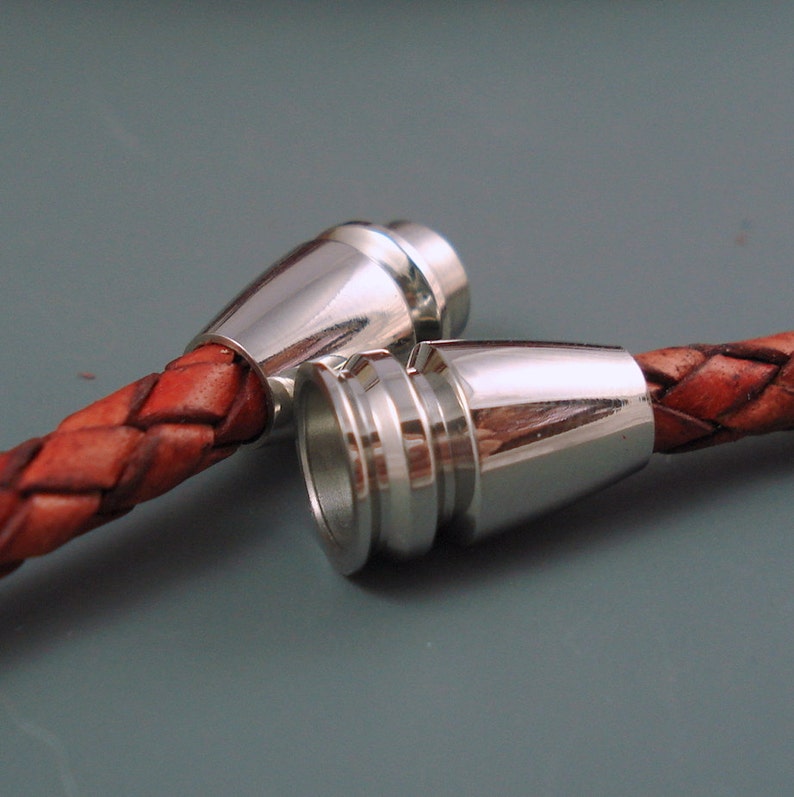 May include: Close-up of two silver-colored magnetic clasps attached to braided brown leather. The clasps have a conical shape and are designed to connect. The leather is a rich, reddish-brown color. The background is a solid, muted gray.