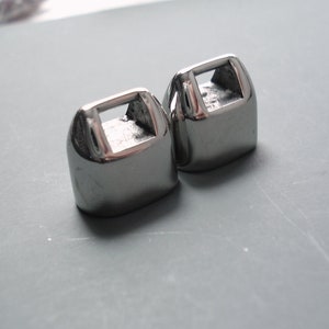 May include: Two silver metal decorative hardware pieces with a square opening in the center. The pieces are shaped like a small house.