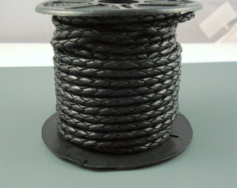 May include: A spool of black braided leather cord. The cord is tightly wound around a black plastic spool. The braided texture is visible, and the cord appears to be new and unused. The spool sits on a gray surface.
