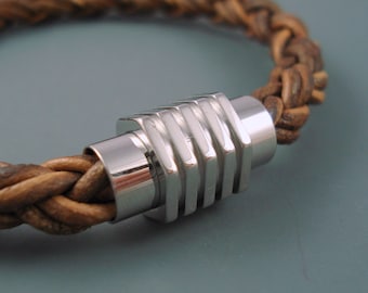 May include: A brown leather braided bracelet with a silver metal clasp. The clasp has a geometric design with multiple ridges.