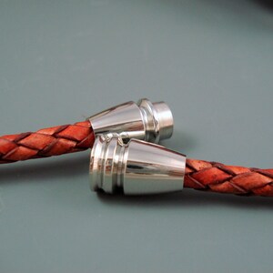 May include: Close-up of two braided leather bracelets with silver-colored magnetic clasps. The leather is a reddish-brown color, and the clasps have a shiny, metallic finish. The bracelets are displayed against a gray background.