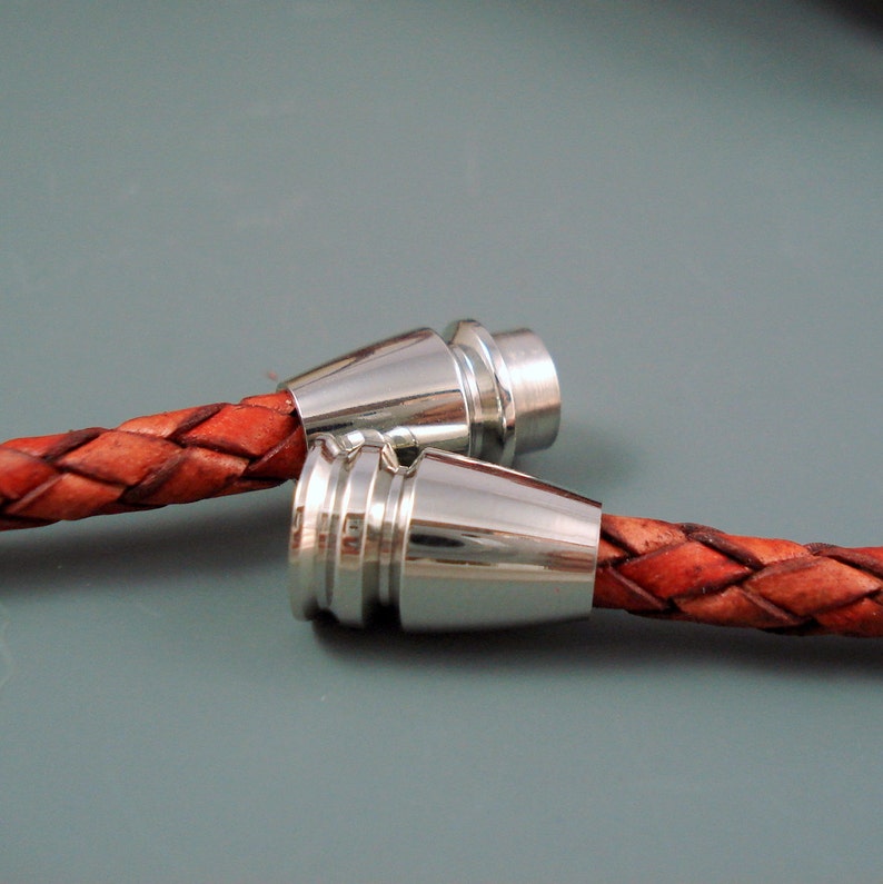 May include: Close-up of two braided leather bracelets with silver-colored magnetic clasps. The leather is a reddish-brown color, and the clasps have a shiny, metallic finish. The bracelets are displayed against a gray background.